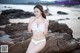 A woman in a white bikini sitting on a rock on the beach.